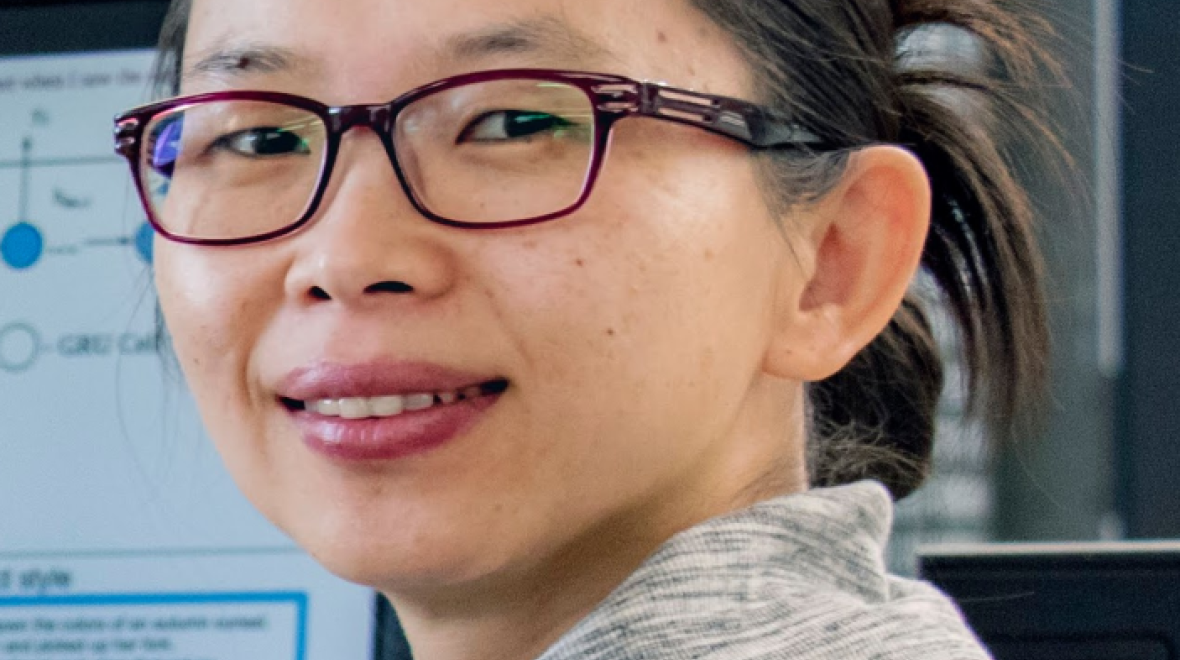 Headshot of Professor Lexing Xie smiling, in the background is a computer screen