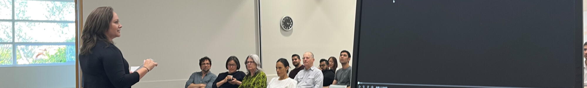 Professor Kate Henne addresses the room of audience. Kate is a woman with long brown hair, wearing khaki skirt and black top. 